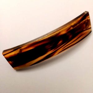 Könnte beinhalten: Eine rechteckige Haarspange mit glänzender, durchscheinender Oberfläche und einem marmorierten Muster in Dunkelbraun- und Bernsteintönen. Das Accessoire hat eine leicht gebogene, längliche Form mit glatten, abgerundeten Kanten.