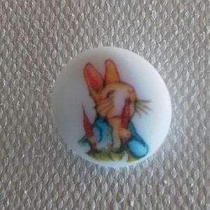 Könnte beinhalten: Weißer Keramikknopf mit einem bunten Bild von Peter Rabbit, der eine blaue Jacke trägt und eine Karotte hält.