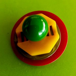 Könnte beinhalten: Vintage Brosche mit mehrschichtigem Design. Eine glänzende grüne Kugel sitzt auf einem gelben, sechseckigen Stück mit Aussparungen, das über einer dunklen Scheibe und einer roten kreisförmigen Basis liegt. Die Brosche befindet sich vor einem grünen Hintergrund.