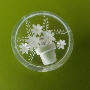 May include: A clear glass paperweight with a white etched design of a potted plant with flowers.