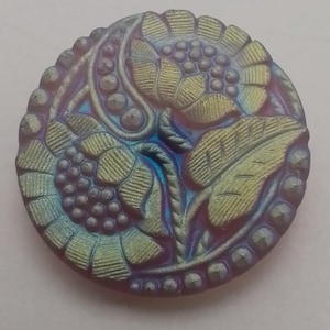 May include: A round iridescent glass button featuring a floral design. The button is a deep purple colour with gold and blue highlights. The design incorporates two stylised flowers with leaves and a beaded border.