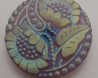 May include: A round iridescent glass button featuring a floral design. The button is a deep purple colour with gold and blue highlights. The design incorporates two stylised flowers with leaves and a beaded border.