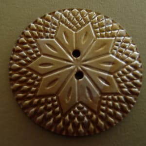 May include: A round, brown button featuring a central eight-pointed star design surrounded by a textured, diamond-patterned border. The button has two small, circular holes positioned vertically in the center for attachment purposes.
