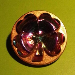 May include: A round, iridescent button with a four-leaf clover design. The clover is multi-colored, reflecting hues of purple, green, and gold. The button has a textured, gold-colored border and sits against a green background.