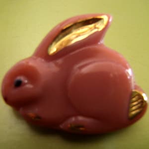 May include: A pink glass rabbit figurine with gold accents. The rabbit has a long, floppy ear and a small, round body.