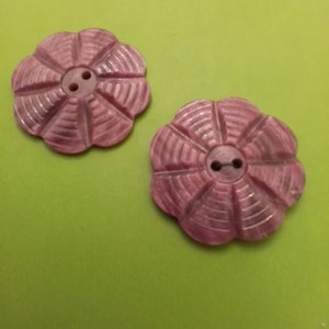 May include: Two pink flower-shaped buttons with a ridged petal design and two small holes for sewing. The buttons are set against a bright green background, showcasing their color and detail.