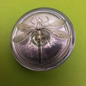 May include: A silver-colored, circular bottle stopper with a detailed dragonfly design. The dragonfly is centered on the stopper, with its wings outstretched. The stopper has a textured, reflective surface, set against a green background.