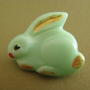 May include: A light green glass rabbit button with gold accents. The rabbit has a pink nose and black eyes.