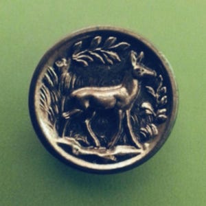 May include: A silver-toned button featuring a detailed image of a deer standing in a field of tall grass. The deer is facing right and has a long, slender neck and legs.