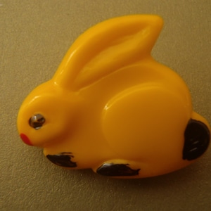 May include: A yellow plastic bunny button with black painted details. The button has a shiny, clear plastic eye.
