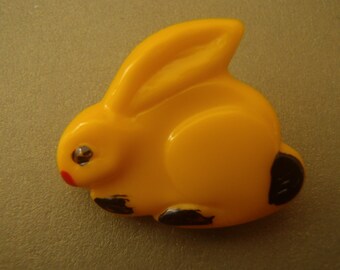 May include: A yellow plastic bunny button with black painted details. The button has a shiny, clear plastic eye.