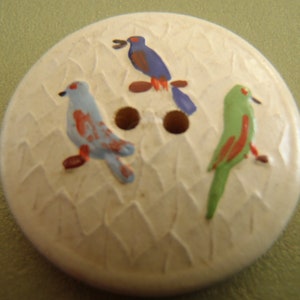 May include: A round, off-white button with a textured surface and three colorful birds. One bird is blue with red accents, another is green with red accents, and the third is purple with orange accents. The button has two brown holes.