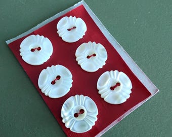 May include: A set of six round, white, iridescent buttons with a carved, scalloped edge and two centre holes, arranged in two columns on a rectangular red card. Each button features a distinct radial pattern radiating from the centre holes.