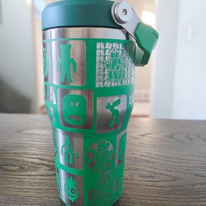 Op de afbeelding: Een groene en zilveren ge&iuml;soleerde beker met een groen deksel en handvat. De beker heeft ge&euml;tste cartoonkarakterontwerpen en het woord "ROBLOX" in een herhalend patroon.