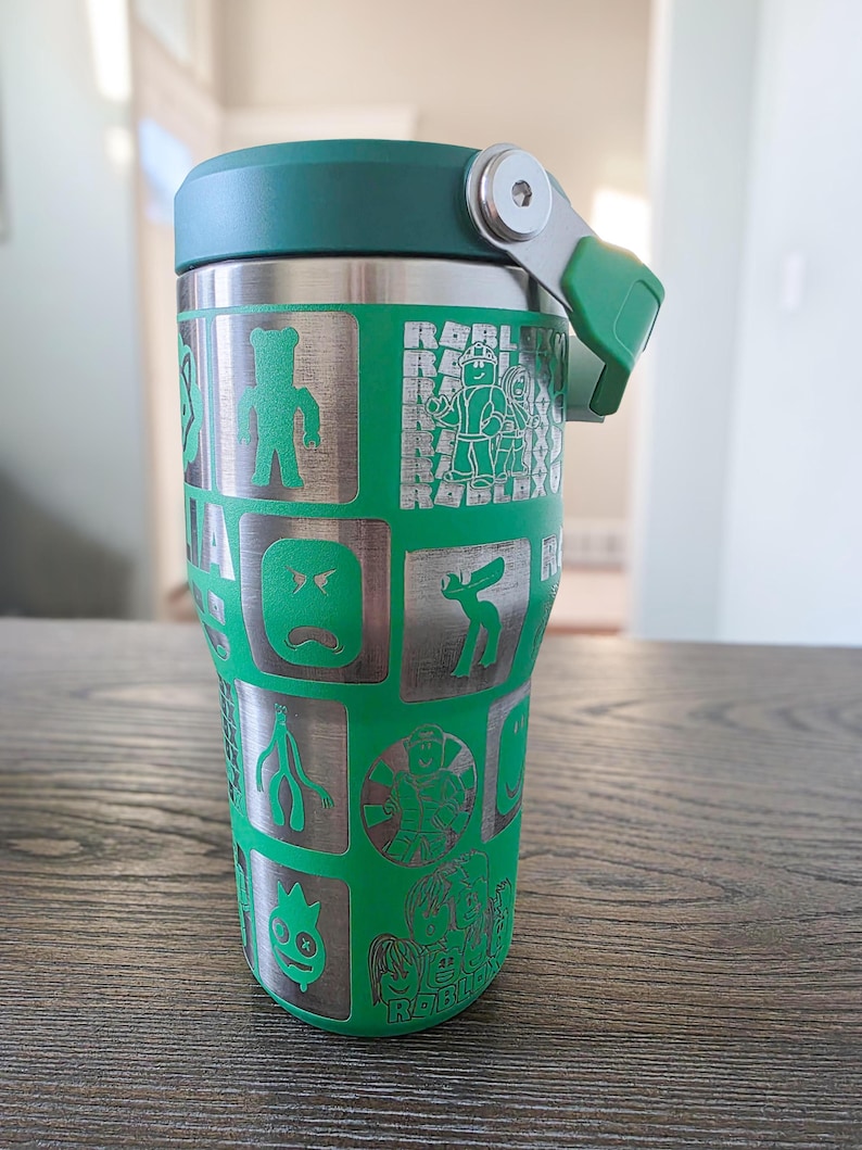 Op de afbeelding: Een groene en zilveren ge&iuml;soleerde beker met een groen deksel en handvat. De beker heeft ge&euml;tste cartoonkarakterontwerpen en het woord "ROBLOX" in een herhalend patroon.
