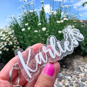 May include: A clear acrylic keychain with the name "Karlie" in white script, accented with glitter. The keychain features a ballet shoe design. The background includes flowers and a blue sky.