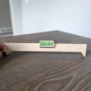 May include: A wooden level tool with a clear, green-filled vial. The level is attached to a light-colored wooden plank. The tool is designed to assist in ensuring surfaces are perfectly horizontal or vertical. The background is a neutral gray.