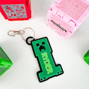 May include: A green and black Minecraft Creeper-shaped keychain with the name "RYDER" in green letters. The keychain has a silver clasp and a black ring. Other colorful toy blocks are in the background.