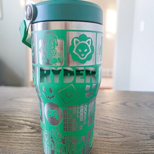 Op de afbeelding: Een groene en zilveren beker met een donkergroen deksel en handvat. De beker heeft ge&euml;tste ontwerpen van videogamepersonages en logo's, met de naam "RYDER" prominent weergegeven. De beker is ontworpen om dranken koud of warm te houden.
