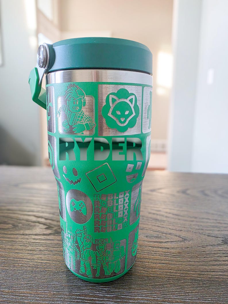 Op de afbeelding: Een groene en zilveren beker met een donkergroen deksel en handvat. De beker heeft ge&euml;tste ontwerpen van videogamepersonages en logo's, met de naam "RYDER" prominent weergegeven. De beker is ontworpen om dranken koud of warm te houden.