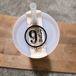 May include: A white tumbler with a straw, featuring a decorative charm with the number "9 3/4" in a black circle. The charm is set against a glittery background with small heart and key accents.