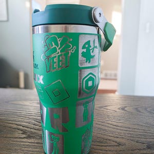 Op de afbeelding: Groene en zilveren ge&iuml;soleerde tumbler met een groen deksel. De tumbler heeft ge&euml;tste ontwerpen van Roblox-gamekarakters en het woord "YEET". De tumbler is ontworpen om dranken koud of warm te houden.