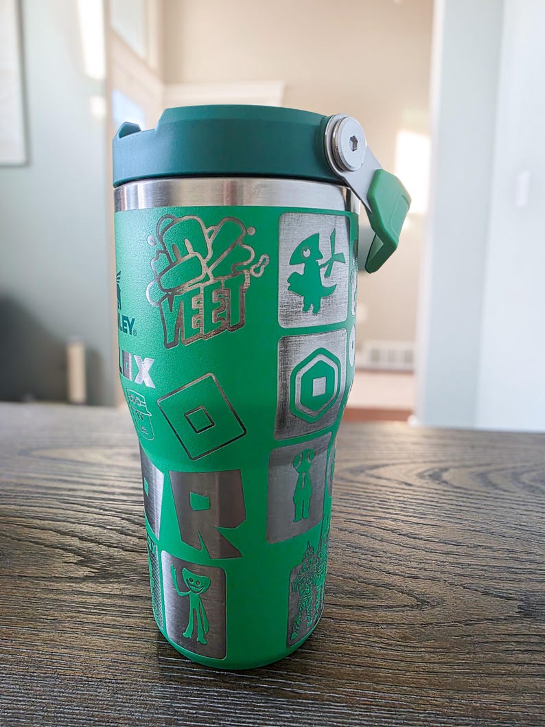 Op de afbeelding: Groene en zilveren ge&iuml;soleerde tumbler met een groen deksel. De tumbler heeft ge&euml;tste ontwerpen van Roblox-gamekarakters en het woord "YEET". De tumbler is ontworpen om dranken koud of warm te houden.