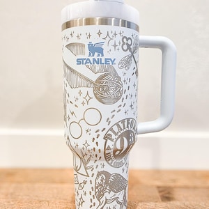 May include: White Stanley tumbler with a handle and straw, featuring a silver design with Harry Potter-themed graphics. The design includes the Stanley logo, Platform 9 3/4, and other iconic imagery. A practical and stylish drinkware item.