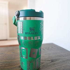 Op de afbeelding: Groene Stanley-beker met een groen deksel en handvat. De beker heeft het Roblox-logo en karakterafbeeldingen. Het Stanley-logo is zichtbaar. De beker staat op een houten oppervlak.