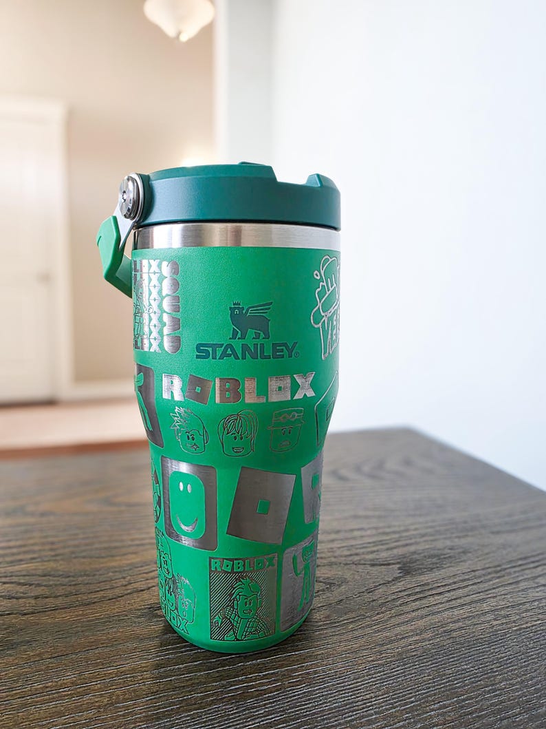 Op de afbeelding: Groene Stanley-beker met een groen deksel en handvat. De beker heeft het Roblox-logo en karakterafbeeldingen. Het Stanley-logo is zichtbaar. De beker staat op een houten oppervlak.