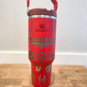 May include: Red Stanley tumbler with a handle and a dark red lid. The tumbler features the text "STANLEY" and "HELLDIVERS II" with various etched symbols. The handle and lid are a darker shade of red.