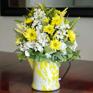 May include: A floral arrangement featuring bright yellow daisies, white blossoms, and green fern foliage displayed in a white pitcher with a yellow marbled pattern and a black handle. The bouquet is dense and rounded, showcasing a mix of textures.