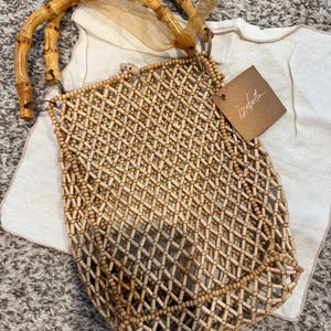 Pu&ograve; includere: Una borsa a mano con perline marrone chiaro con manico in bamb&ugrave;. La borsa ha un motivo intrecciato e un'etichetta con la scritta "Izabella". La borsa &egrave; appoggiata su un panno bianco.