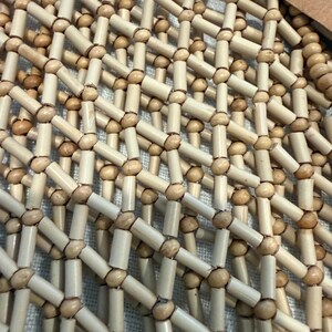 Pu&ograve; includere: Primo piano di una borsa o stuoia tessuta con perline di legno e pezzi cilindrici di colore naturale. Il design presenta un motivo a griglia con piccole perline rotonde alle intersezioni. La trama appare liscia e il colore &egrave; beige chiaro.