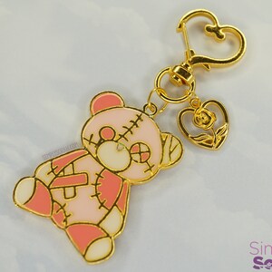 May include: A pink and white stitched teddy bear charm with a gold heart charm featuring a rose. The charms are attached to a gold key chain.