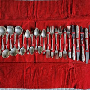 May include: A set of silver-toned flatware arranged in a red fabric roll. The collection includes various spoons, forks, and knives, each featuring decorative embossed patterns on the handles. The utensils are held in place by red elastic loops.
