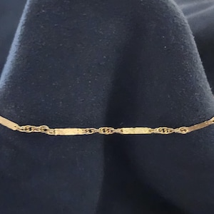 May include: A delicate gold chain bracelet featuring a repeating pattern of twisted rope links alternating with smooth, flat rectangular bar links. The metal has a polished finish that reflects light along the entire length of the jewelry piece.