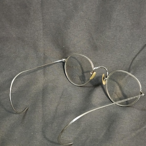 May include: A pair of silver-toned metal eyeglasses featuring round frames and thin, curved temple arms. The glasses include small, pale yellow nose pads attached to the bridge, providing a clear view of the circular lenses and the metallic structure.