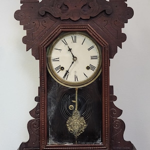 May include: Antique wooden clock with a white clock face and black Roman numerals. The clock has a pendulum and a decorative brass-colored weight. The wooden frame is dark brown with ornate carvings.