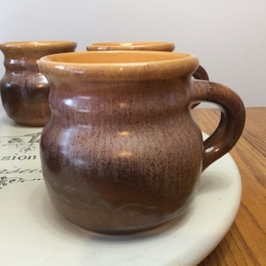 Puede incluir: Conjunto de tres tazas de cerámica marrón y amarillo con un esmalte moteado. Las tazas tienen un cuerpo redondeado y un asa.