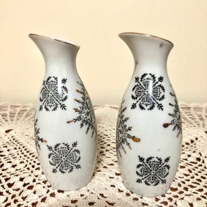 May include: Two white ceramic vases with black and gold geometric designs. The vases are on a white crocheted surface.