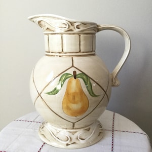 May include: A white ceramic pitcher with a pear design. The pitcher has a round body with a decorative base and a handle. The pear is painted in shades of yellow, green, and brown.