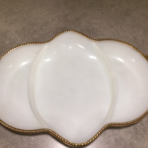 May include: A white, three-section serving dish with a gold-colored beaded trim. The dish has a unique, scalloped shape and is likely made of glass or ceramic. The gold trim adds a touch of elegance to the serving piece.