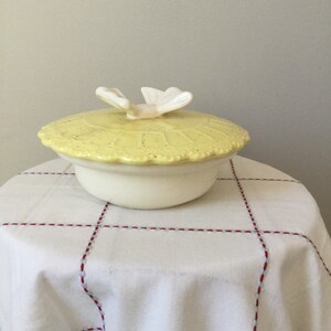 Puede incluir: Un recipiente de cerámica amarillo con una tapa de mariposa blanca. El recipiente tiene una base blanca y una tapa amarilla con un diseño en relieve. La tapa tiene una mariposa blanca en la parte superior.