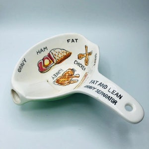 Puede incluir: Separador de salsa de cerámica blanca con un borde verde. El separador tiene ilustraciones de diferentes carnes, incluyendo jamón, pavo, pollo y grasa. El mango está etiquetado como "Separador de salsa grasa y magra".
