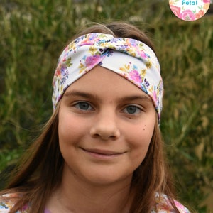 Puede incluir: Una persona joven lleva una diadema blanca con un estampado floral. La diadema está anudada en la parte superior de la cabeza. La persona lleva una camisa blanca con un estampado floral. La camisa tiene un ribete lavanda alrededor del cuello. La persona tiene el pelo largo y castaño.