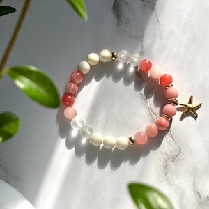 May include: A pink and white beaded bracelet with a gold starfish charm. The bracelet is made with natural stones and is perfect for layering.