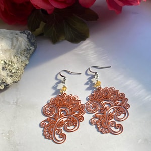 Peut inclure: Une paire de boucles d'oreilles pendantes avec un motif de filigrane de couleur orange brûlée. Les boucles d'oreilles ont un crochet argenté et une petite perle transparente suspendue au sommet.