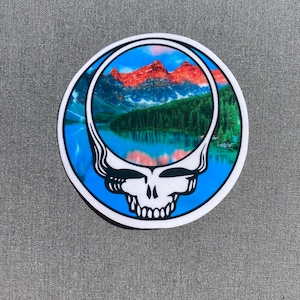 May include: A round sticker with a blue, white, and red tie-dye design. The center of the sticker features a mountain lake scene with a mountain range in the background. The sticker is a parody of the Grateful Dead's iconic Steal Your Face logo.