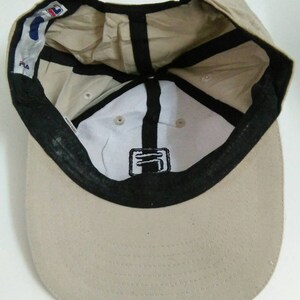 May include: A beige baseball cap with a black brim and a black Fila logo on the front. The cap has a black adjustable strap in the back.